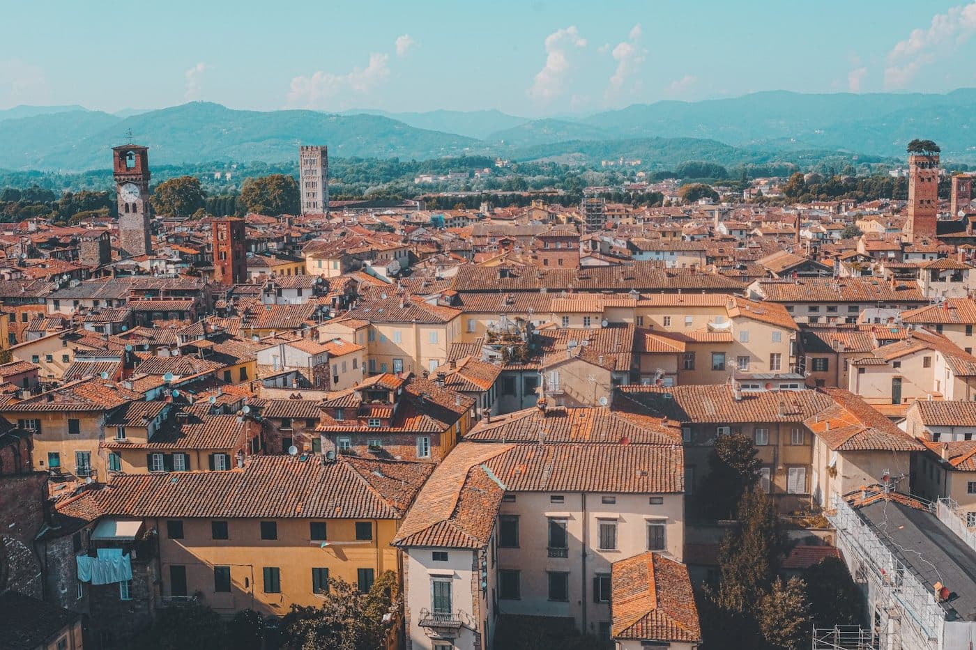 The terracotta rooftops of Lucca from above, with the tree-topped Guinigi tower on the right and mountains behind
