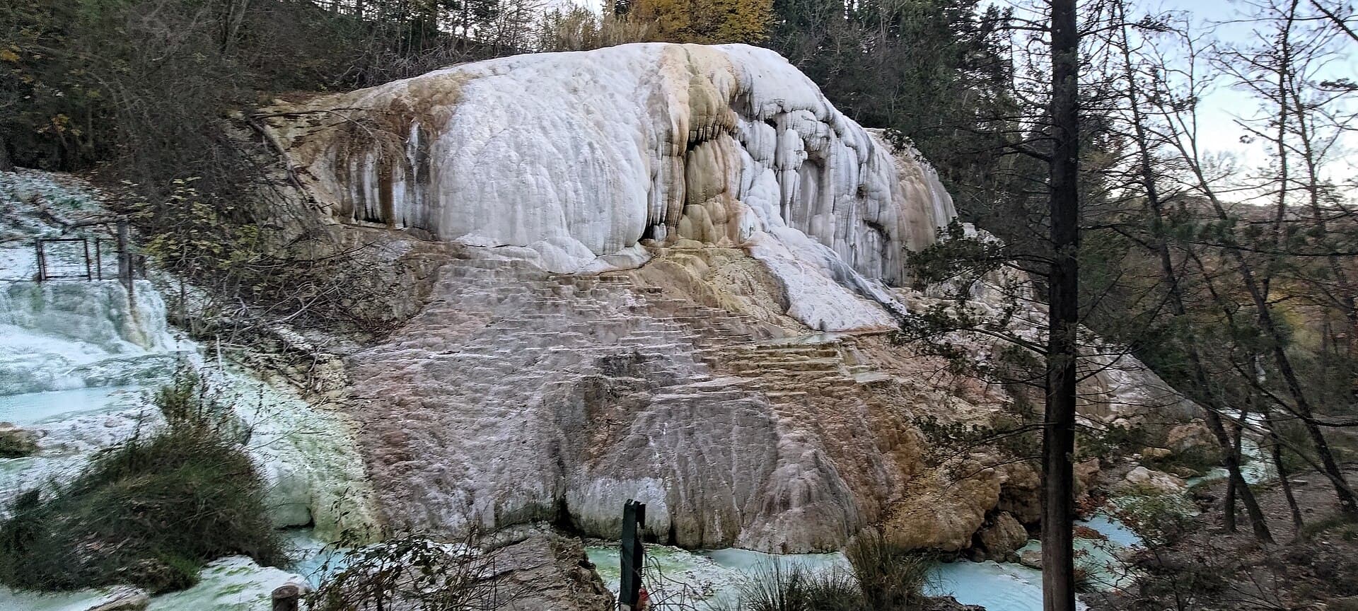 The Balena Bianca at Bagni San Filippo, a large white calcareous formation like a frozen waterfall in autumn woodland, with pale blue water pooling at its foot