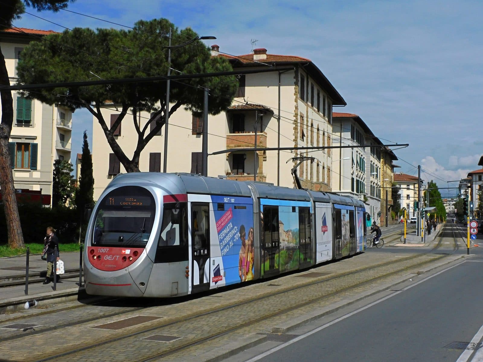 A Florence tram on line T1 wrapped in advertising for the Barberino Designer Outlet reading Fermata Barberino, fino al meno 70 per cento, stopped in a residential street with umbrella pines and shuttered apartment blocks behind it