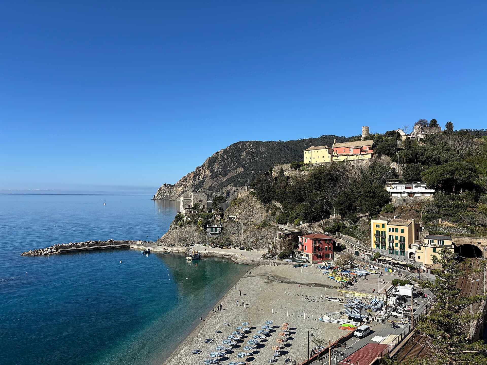 The beach at Monterosso al Mare with rows of parasols, the breakwater, and a railway tunnel mouth at the right of the seafront