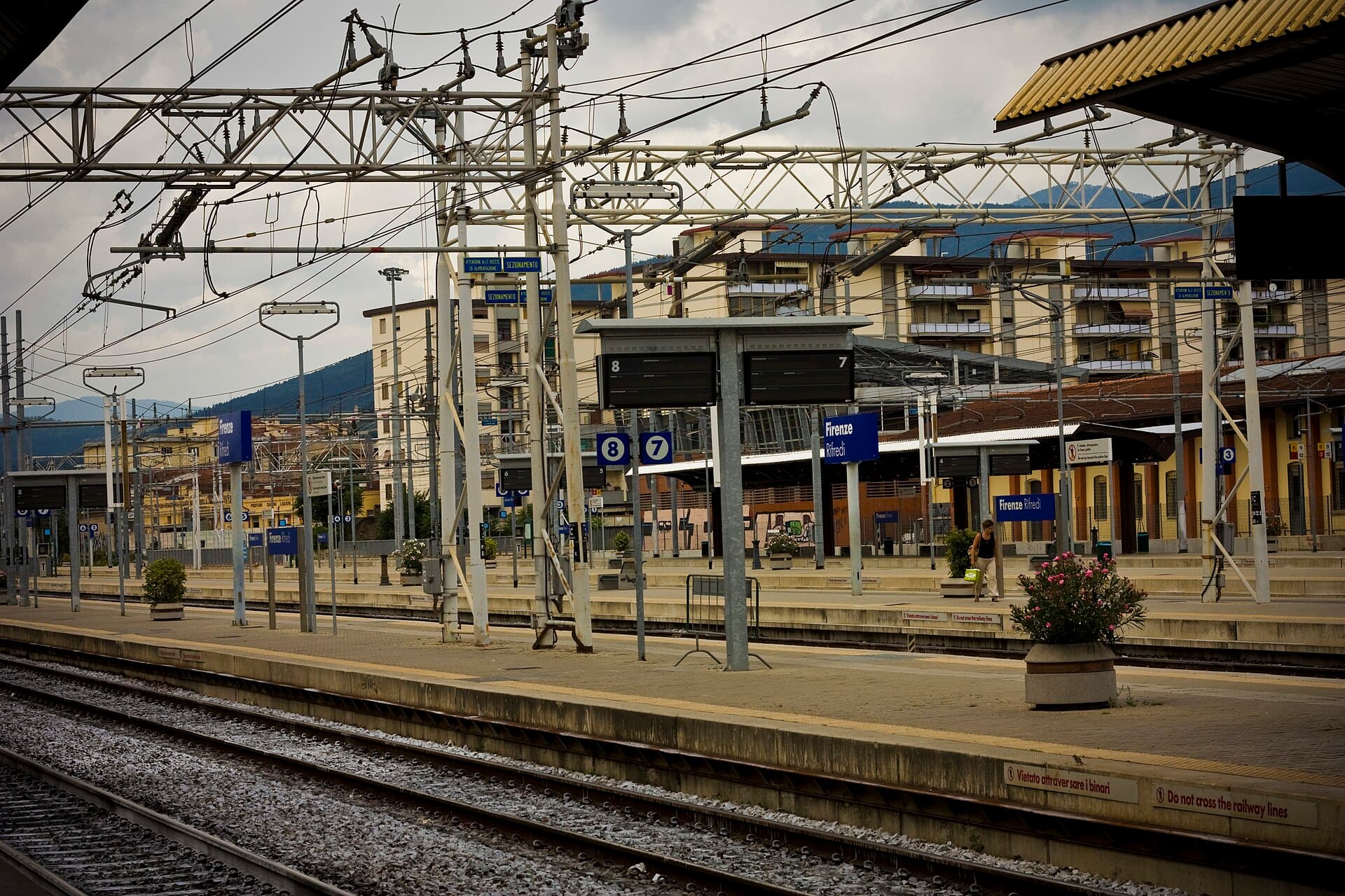 Through platforms at Firenze Rifredi with overhead wires, platform 7 and 8 signs, one traveller walking with a shopping bag and ordinary apartment blocks and hills behind