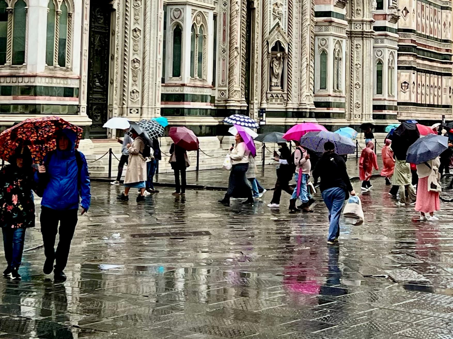 Wet paving outside Florence cathedral with a crowd of people crossing under open umbrellas, most of them in light raincoats rather than heavy winter coats, the marble facade of the Duomo behind them