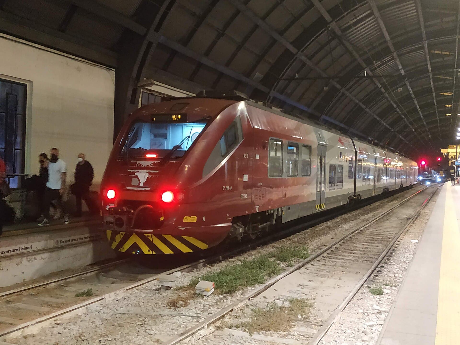 A red Malpensa Express train waiting at a platform inside Milano Centrale station at night, with passengers walking alongside