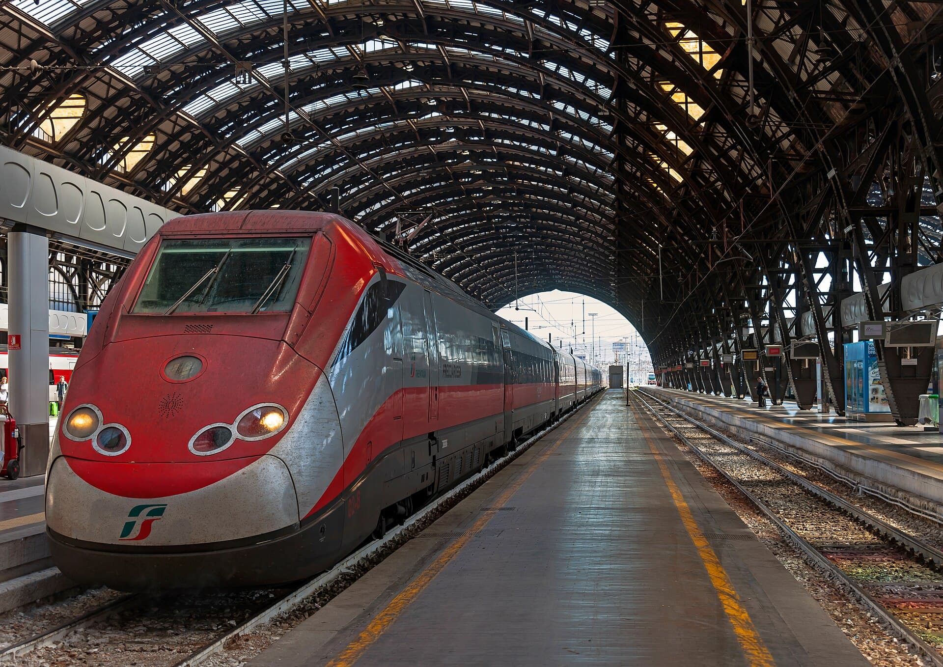 A red and grey Frecciarossa high-speed train waiting at a platform inside Milano Centrale, under the station's arched iron and glass roof