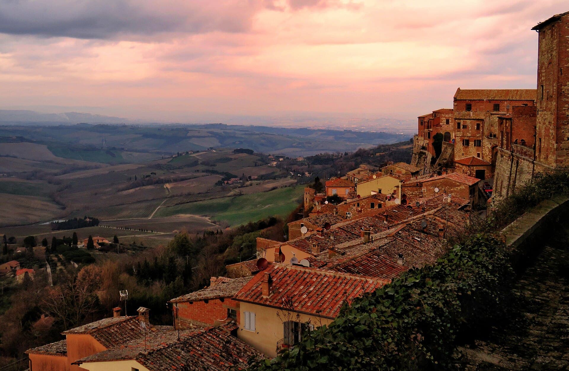 Terracotta rooftops of Montepulciano descending a ridge at sunset, with brick palazzi on the right and the wide valley of the Val d'Orcia below under a pink sky
