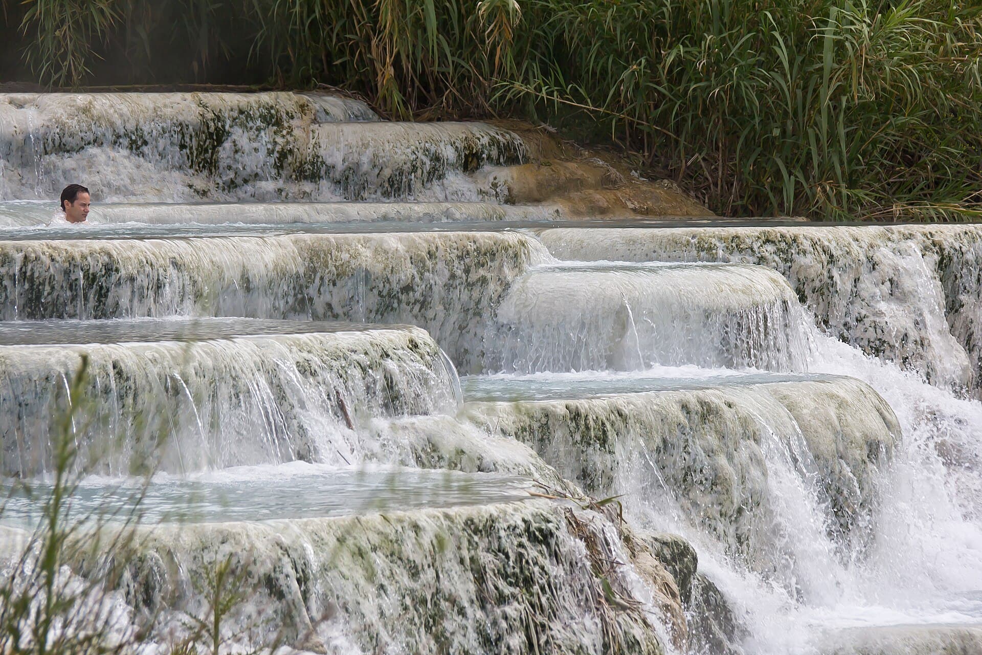 Tiered white travertine terraces at the Cascate del Mulino with hot water spilling from one shallow pool into the next, reeds behind and a single bather visible in an upper pool