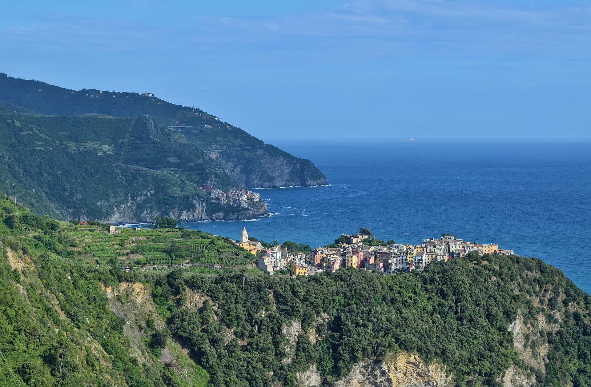 Corniglia seen from the Sentiero Azzurro, a cluster of pastel houses on a headland above terraced vineyards, with Manarola further along the cliffs in the distance