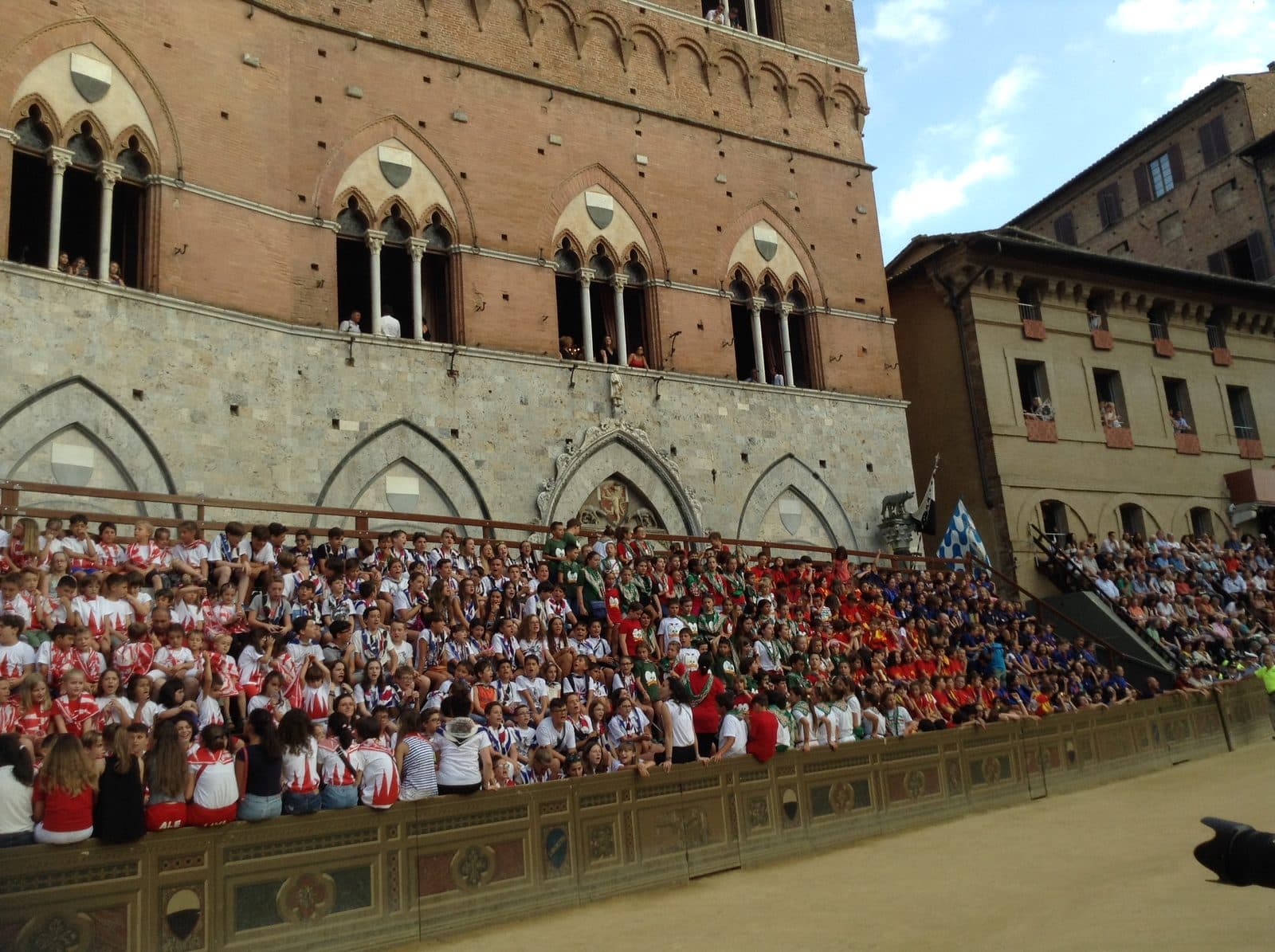 Children in contrada colours packed onto the stands beside the track in Piazza del Campo at a Palio trial, with spectators at the windows and balconies of the Palazzo Pubblico above them and the empty earth track in front